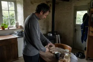 Man in a modest rural kitchen, preparing bread and coffee by a window with subdued natural daylight, running shoes and jacket by a door in the background.