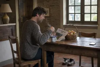 Middle-aged man in a Norman farmhouse kitchen, sitting at a wooden table with coffee, local newspaper, apples, and running shoes nearby, in soft natural morning light.