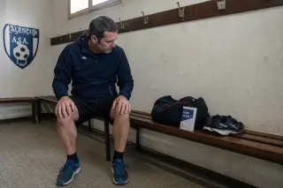 Man in a local Normandy locker room sitting on a wooden bench, looking thoughtfully at an osteopathy info leaflet beside his gym bag and running shoes, in soft natural light.