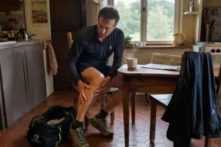 Middle-aged man in a Norman kitchen massaging his calf after a run, surrounded by hints of local everyday life and sports gear.