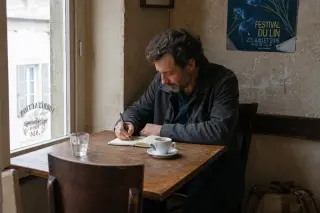 Man in his forties writing in a notebook at a plain wooden table in a rustic Norman café, morning light softly illuminating the calm scene.