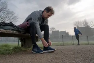 Man tying trail shoes on a bench in a Normandy park at dawn, with another stretching quietly nearby and a yoga mat in a sports bag, soft grey light and local houses in the background.