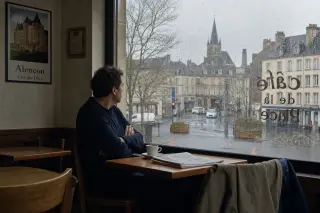 Man in his forties sits alone at a wooden table in a modest café in Alençon, gazing out a rain-streaked window with a cup of coffee, subtle signs of local life and a calm, subdued atmosphere.