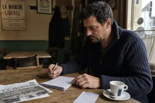 Man in his forties in a Norman village café, reviewing a handwritten checklist with a coffee, in soft natural light, appearing thoughtful and relaxed